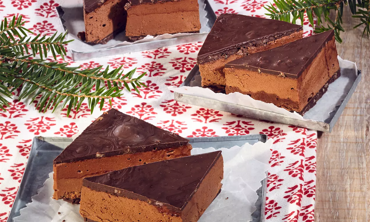 Háromszög alakú nugátos csokoládétrüffel tortaszeletek sötét csokimázzal bevonva, réteges textúrával, fényes tálcákon, fenyőágakkal díszítve