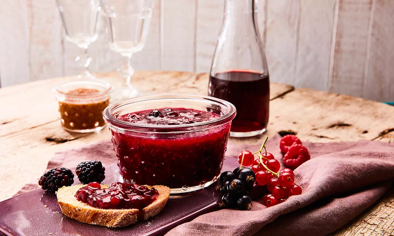Sötét rubinpiros bogyós gyümölcsös chutney üvegtálban, rusztikus faasztalon, mellette kenyérszelet és friss gyümölcsök lazán elrendezve