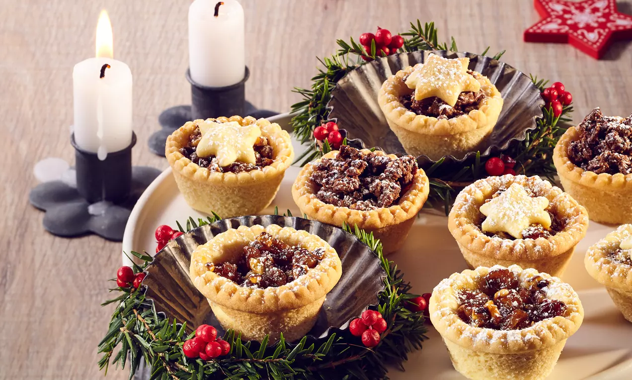 Vajas tésztából készült mini tortácskák hullámos széllel, sötétbarna gyümölcsös töltelékkel, csillag alakú díszekkel, fenyőággal és piros bogyókkal körítve